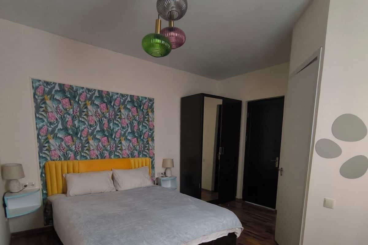 Un dormitorio con una cama, dos mesitas de noche, un armario y una puerta. En la pared, un estampado floral.