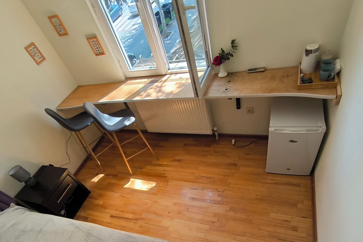 Una habitación pequeña con una cama, un escritorio con silla, un radiador, un refrigerador compacto y una ventana con vistas a coches estacionados.