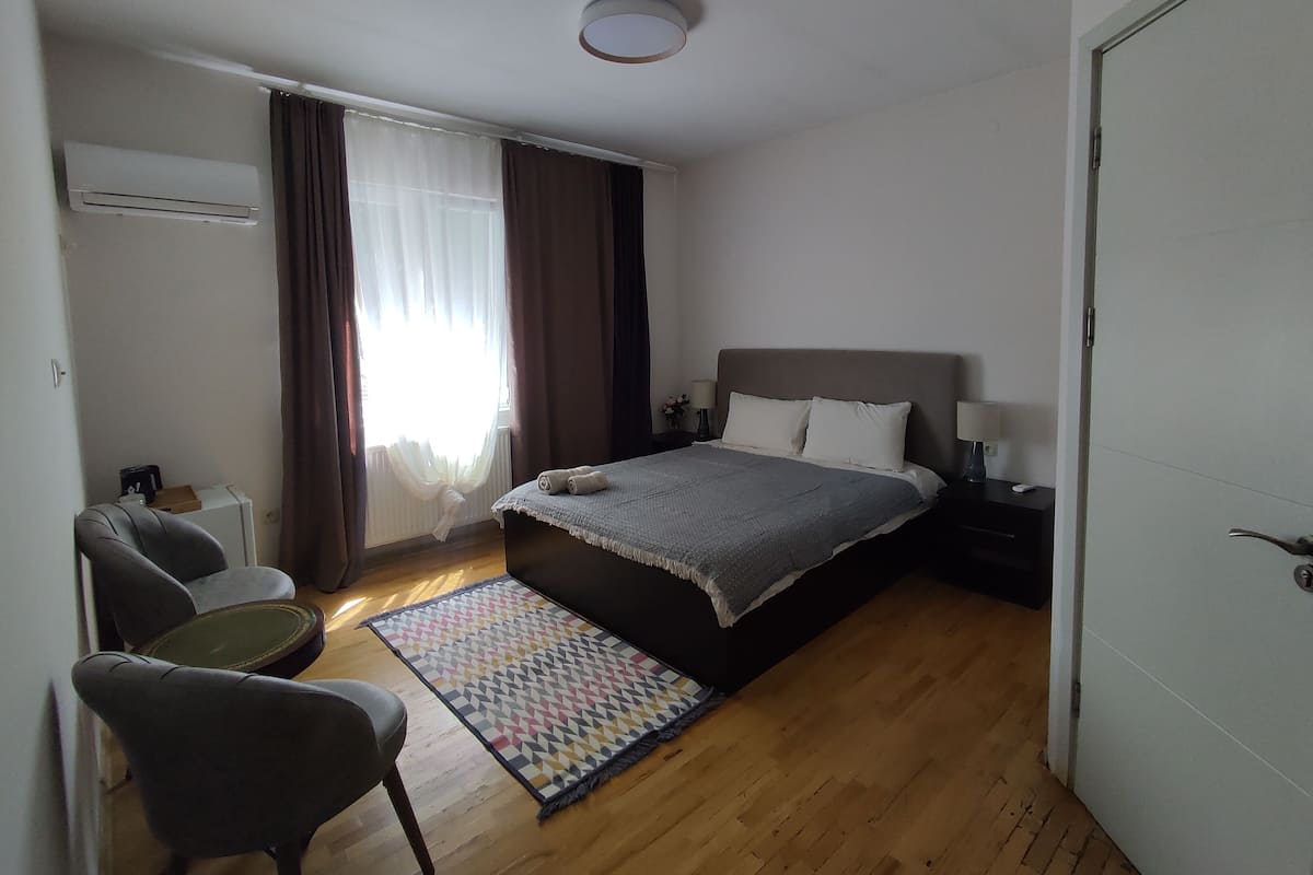 Un dormitorio con cama, mesitas de noche, una silla, alfombra y ventana con cortinas.