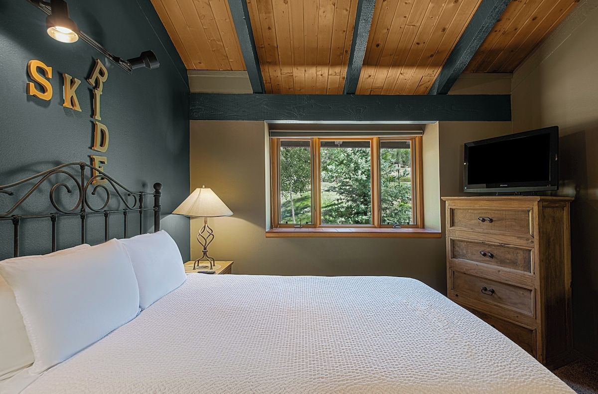 A bedroom with a bed, a nightstand with a lamp, a dresser, a TV, and a window with a view of trees.