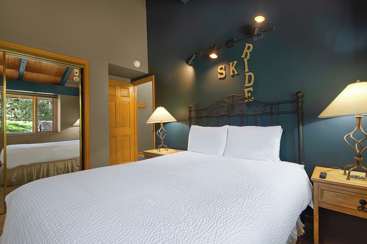 Una cama bien tendida con ropa blanca, dos lámparas de noche y una pared con las palabras 'SKID SKIDE' iluminadas.