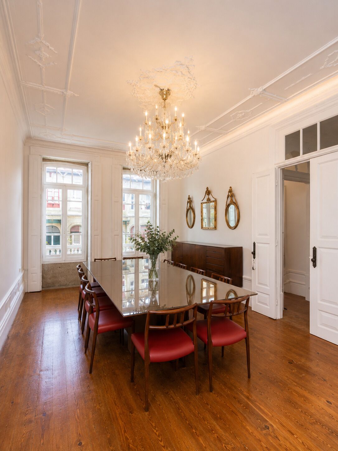 A dining room with a chandelier, wooden dining table, red chairs, and a vase with flowers.