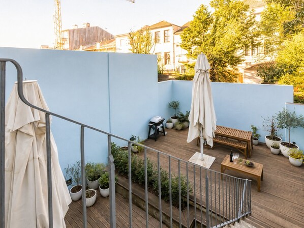 Una terraza en la azotea con piso de madera, mobiliario exterior, plantas en macetas y vistas de edificios circundantes.