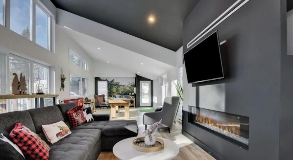 A modern living room with a fireplace, a flat-screen TV, and a dining area visible through an open door.