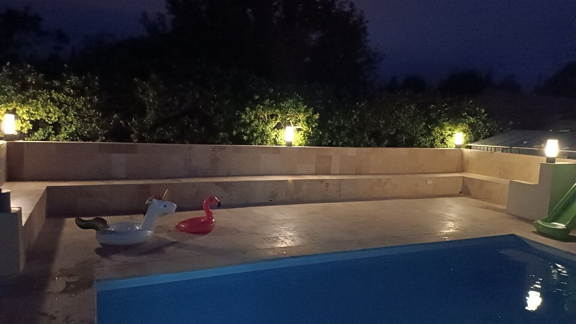 Une piscine agrémentée d’un flotteur en forme de flamant rose, aux couleurs blanche et rouge, entourée d’un petit muret en béton et éclairée par des lampes fixées au mur.