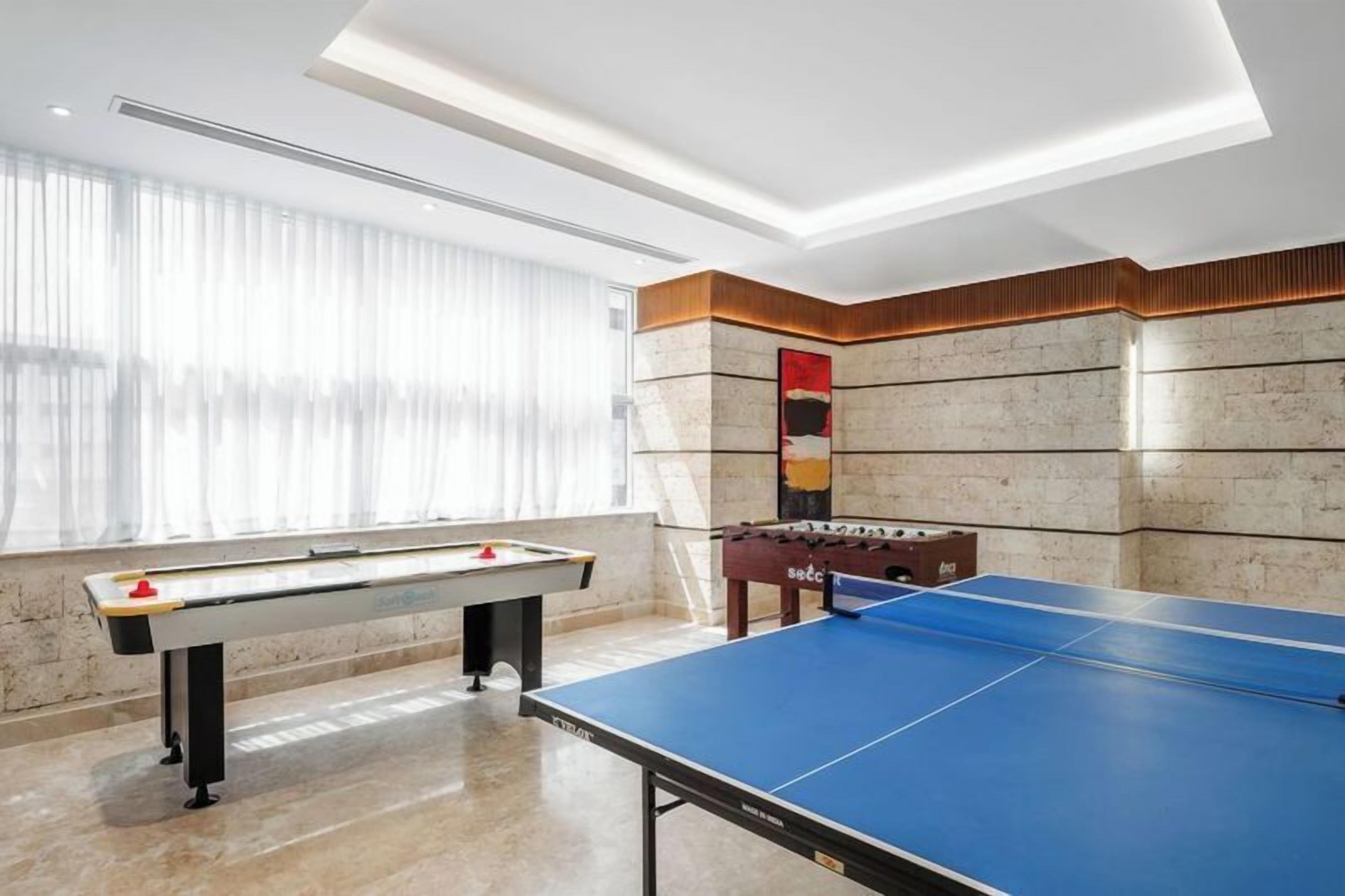A room with a foosball table, a ping pong table, and a window with sheer curtains.