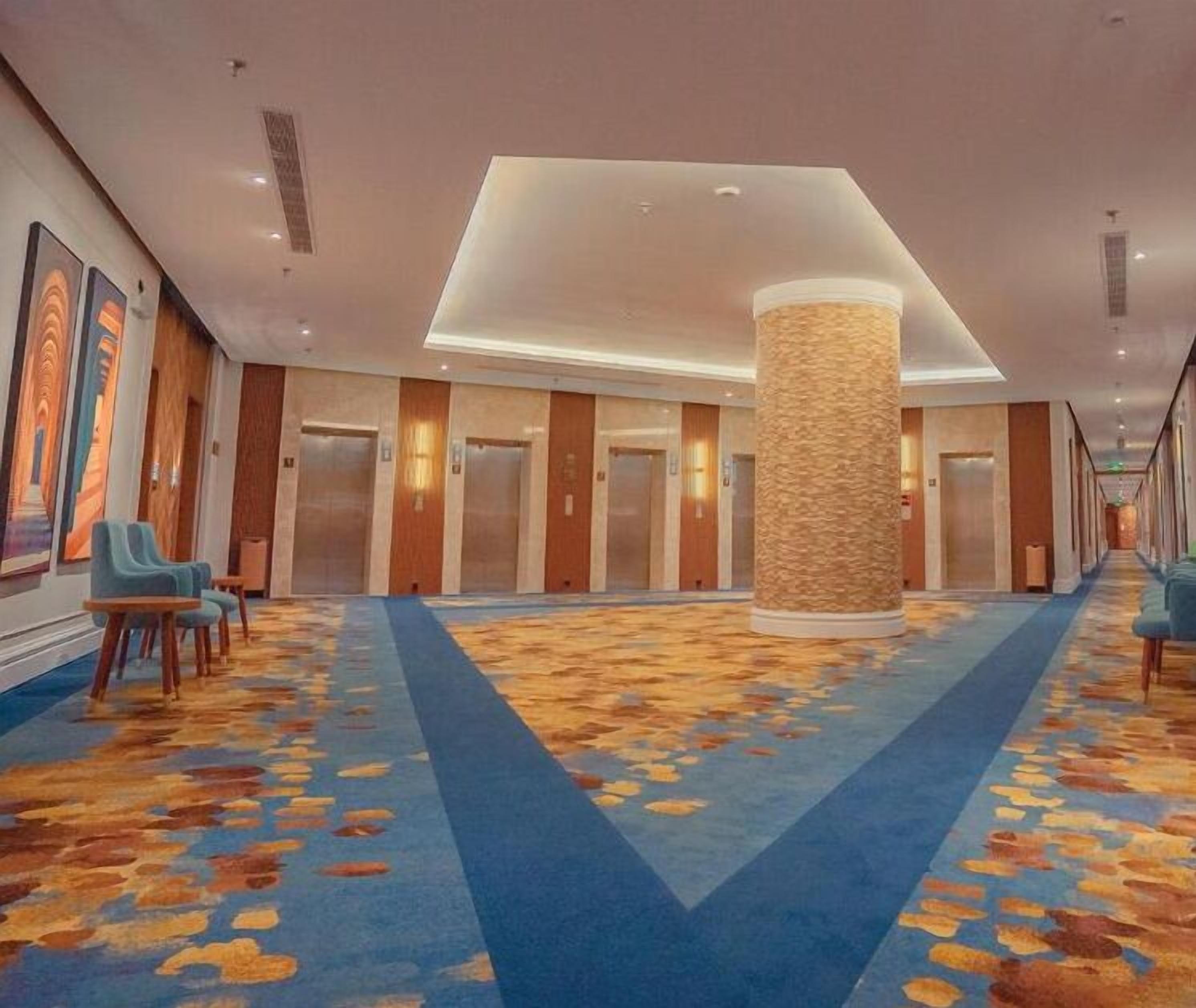 A hotel lobby with a patterned carpet, a central column, and a row of elevators.