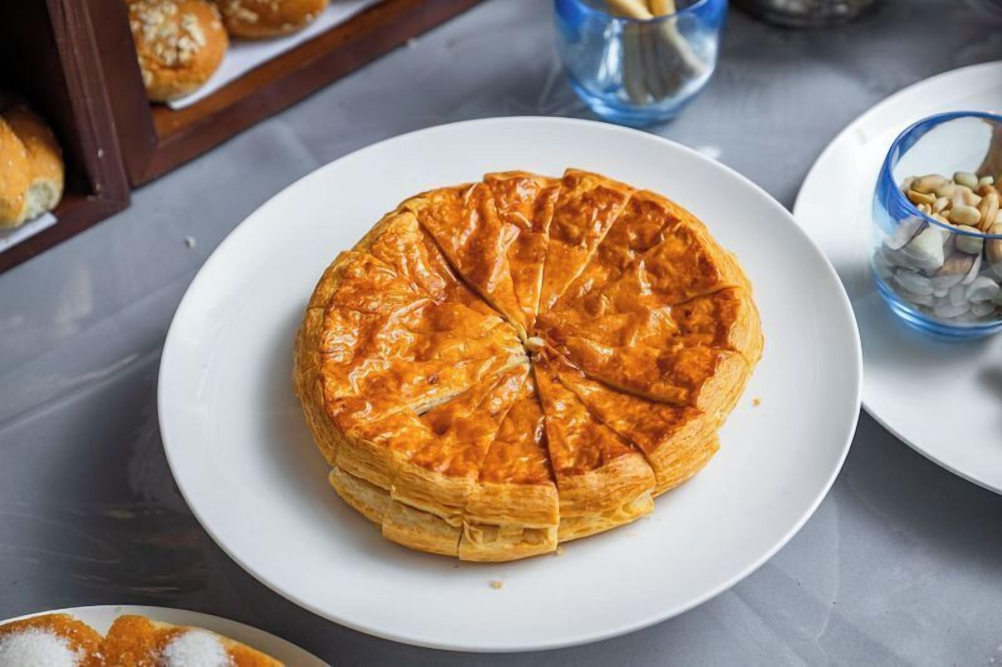 A round pastry on a white plate, with a blue glass and a bowl of nuts in the background.