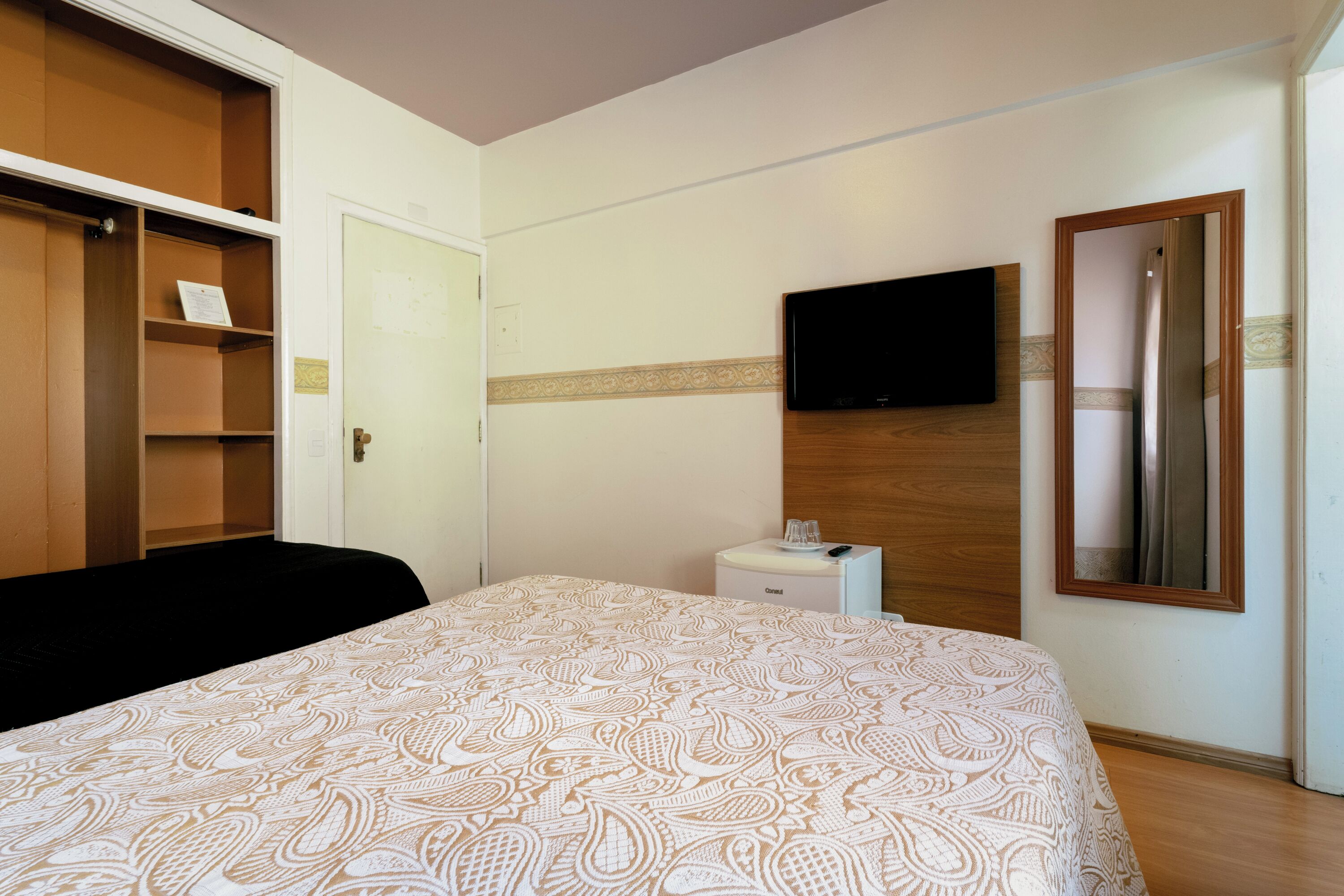 A hotel room with a bed, a wardrobe, a bedside table with a glass, and a mounted TV.