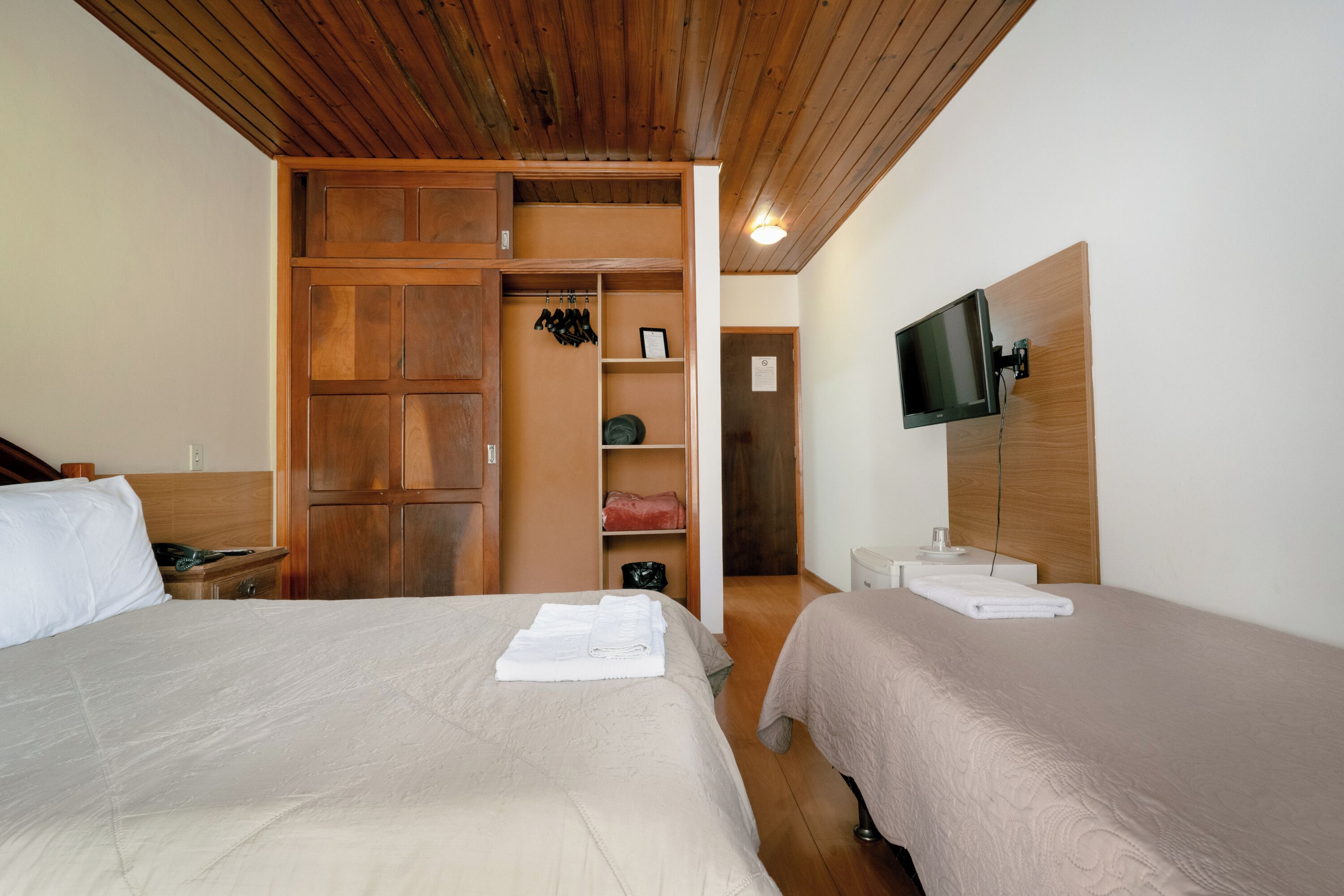 A hotel room with two beds, wooden wardrobe, a television mounted on the wall, and a small table with a towel.