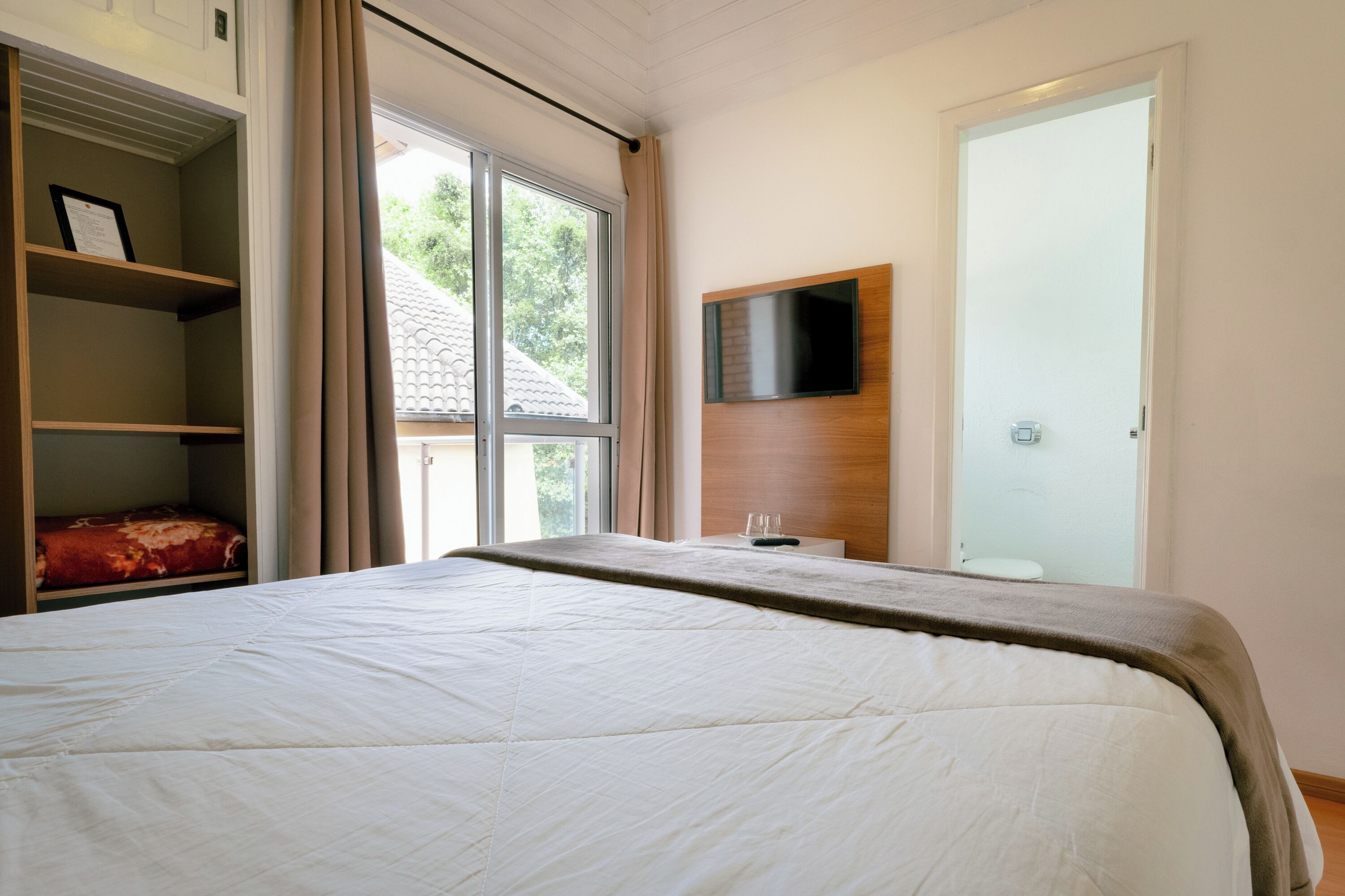 A bedroom with a large bed, a TV mounted on the wall, and a window with curtains.