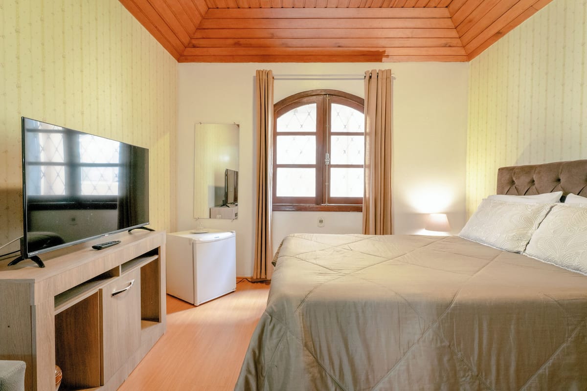 Un dormitorio con una cama, televisión, mesita de noche pequeña, espejo y ventana con cortinas.
