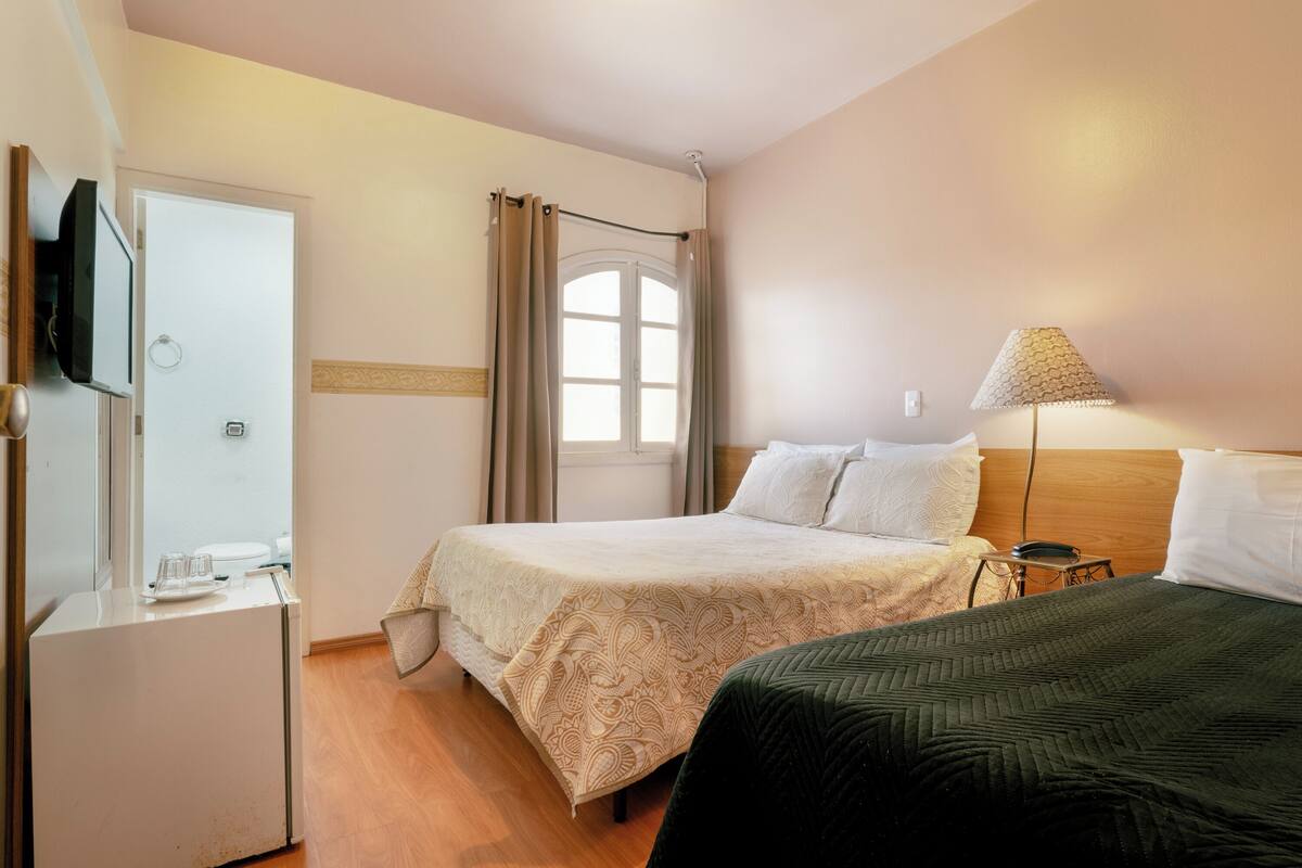 Un dormitorio con una cama, televisión, ventana con cortinas y una lámpara.