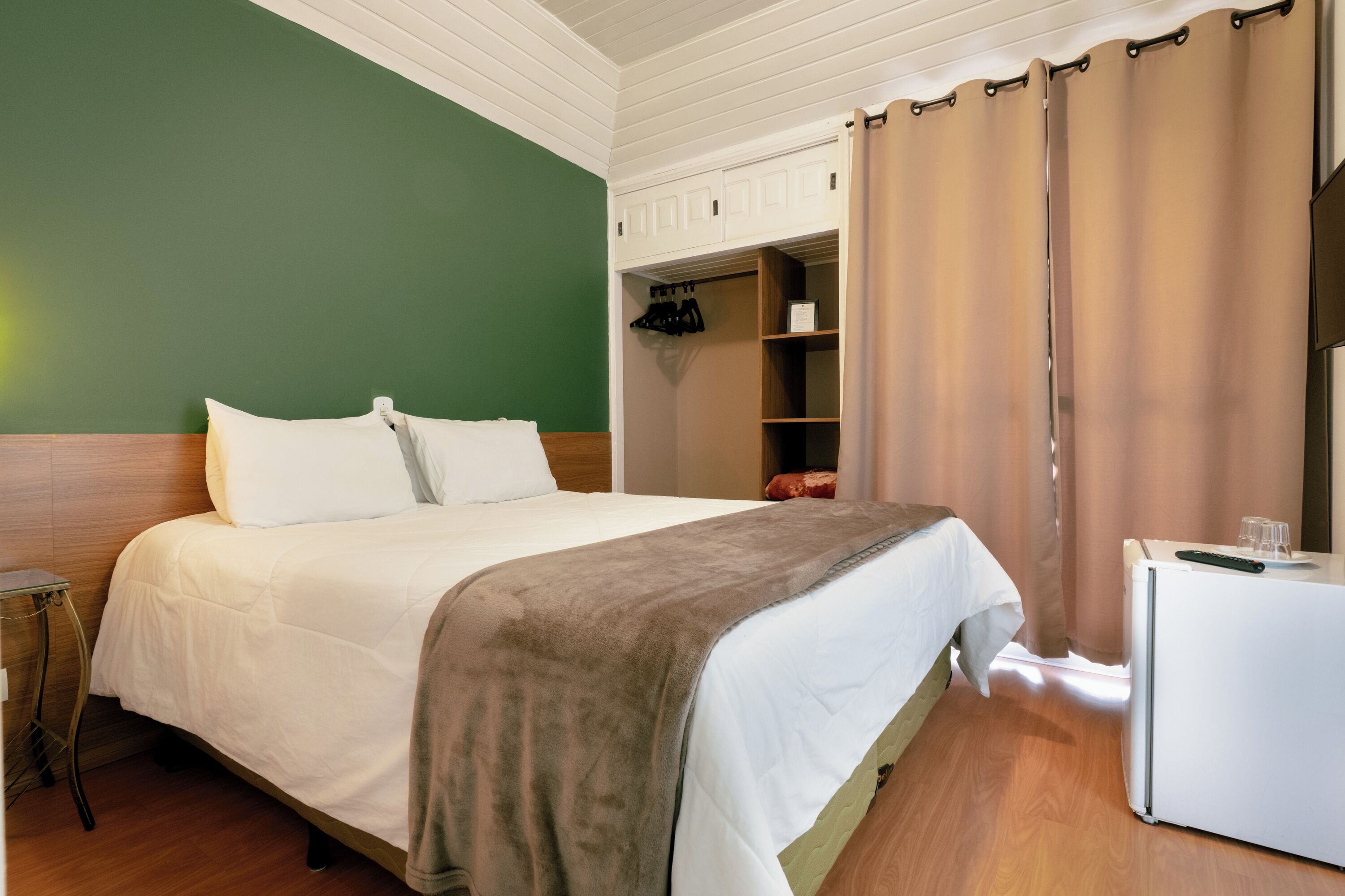 A neatly made bed with a white comforter and a brown throw blanket, a small bedside table, a white wardrobe with hanging space, and a white refrigerator.
