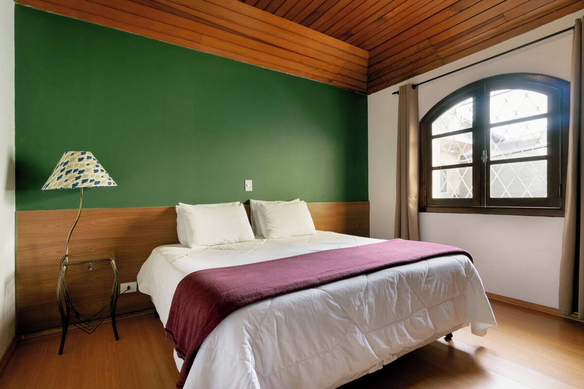 Una cama bien hecha con edredón blanco y manta color borgoña, una lámpara con pantalla estampada y una ventana con marco de madera y cortinas.