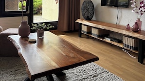 Una sala de estar moderna con una mesa de centro de madera, un televisor de pantalla plana montado en la pared y un amplio ventanal corredero que da a un balcón con vistas a la vegetación y montañas.