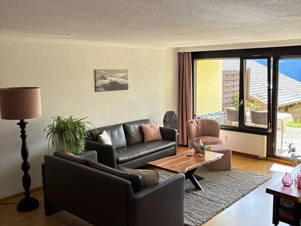 A modern living room with a black leather sofa, a wooden coffee table, a floor lamp, and a large window offering a view of a garden and mountains. - Apartment with great view in a 4* hotel with Pool, Sauna and Fitness (Beatenberg)