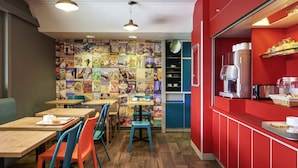 A café with wooden tables and colorful chairs, a red counter with a coffee machine, and a wall decorated with travel posters. - Hotelf1 Marseille Plan De Campagne N°1 (Rénové) (Les Pennes-Mirabeau)