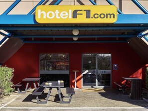 A hotel entrance with a red exterior, a yellow sign reading 'hotelFL.com', and a covered outdoor seating area with tables and benches. - Hotelf1 Marseille Plan De Campagne N°1 (Rénové) (Les Pennes-Mirabeau)