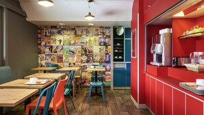 A café with wooden tables and colorful chairs, a red counter with a coffee machine, and a wall decorated with travel posters.