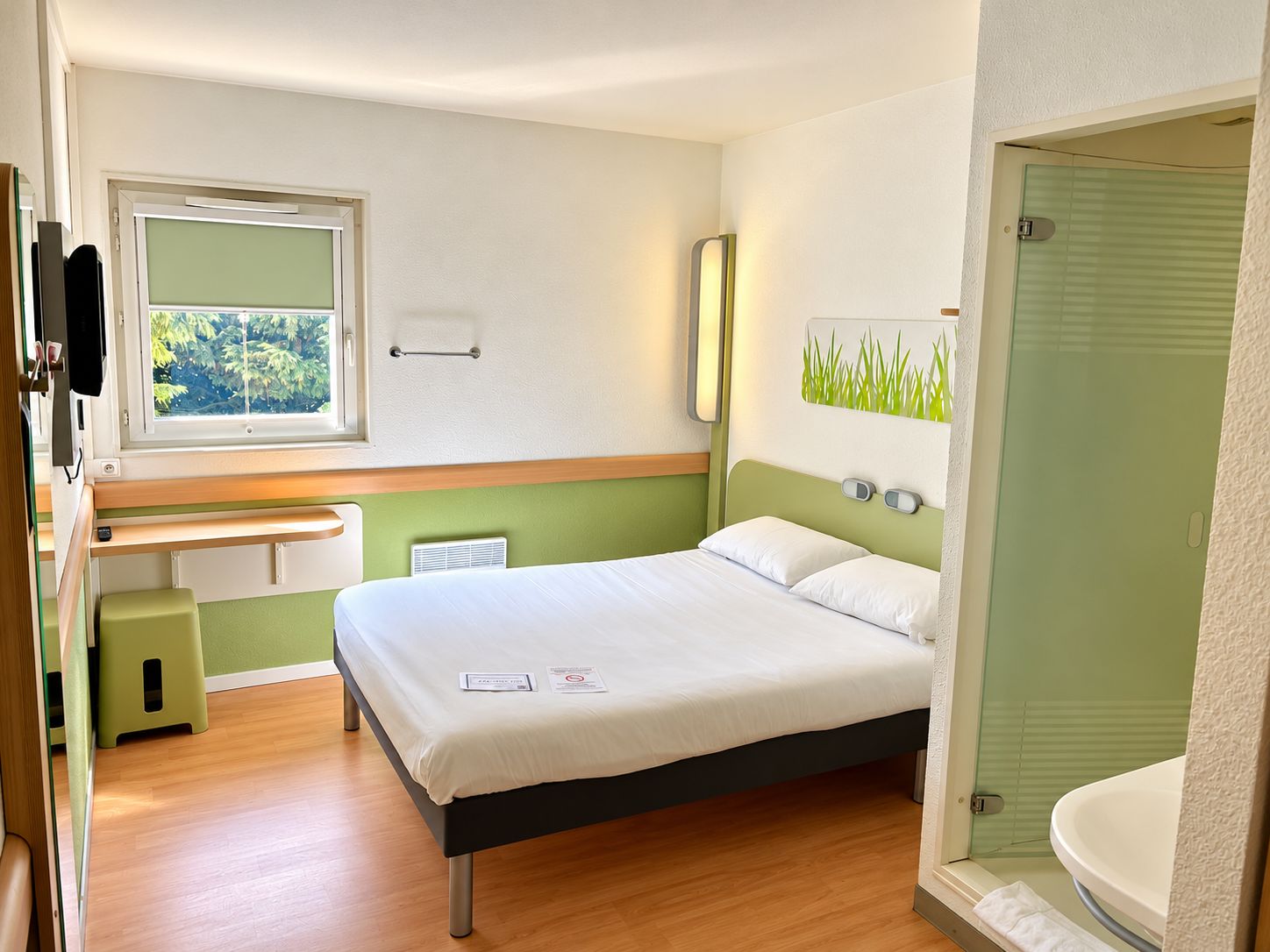 A hotel room with a bed, a bedside table, a mirror, a window with a view of trees, and a bathroom with a sink and toilet.