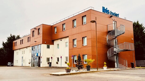 A modern building with a sign reading "ibis budget". It features a corner entrance, multiple windows, and a spiral staircase on the right side. - Ibis Budget Roye (Roye)