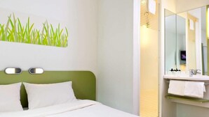 A hotel room with a bed, a green headboard, a wall-mounted mirror, and a bathroom with a sink and towel rack. - Ibis Budget Roye (Roye)