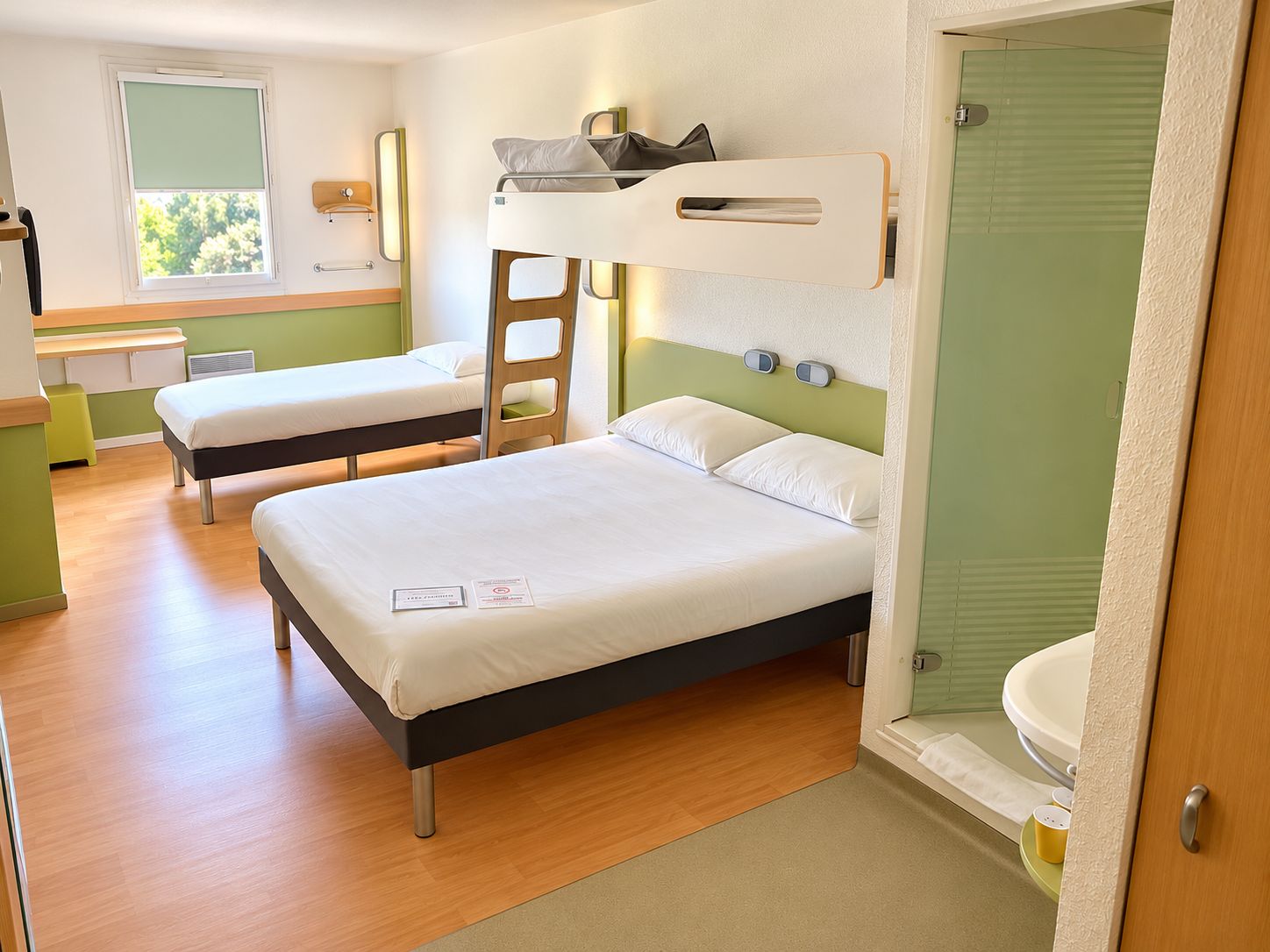 A hotel room with two single beds, a bunk bed, a small desk, a window, and a bathroom.