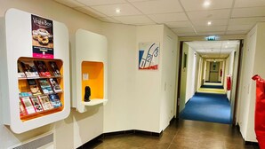 A hotel corridor with a vending machine, a poster, and a fire exit sign. - Ibis Budget Roye (Roye)
