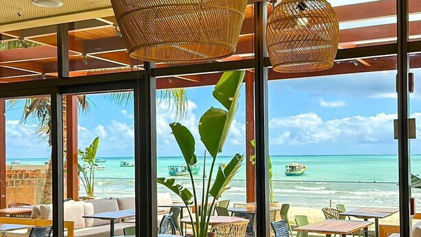 A beachside restaurant with wicker hanging lamps, blue cushioned seating, and large windows offering ocean views. - Ibis Styles Maragogi (Maragogi)
