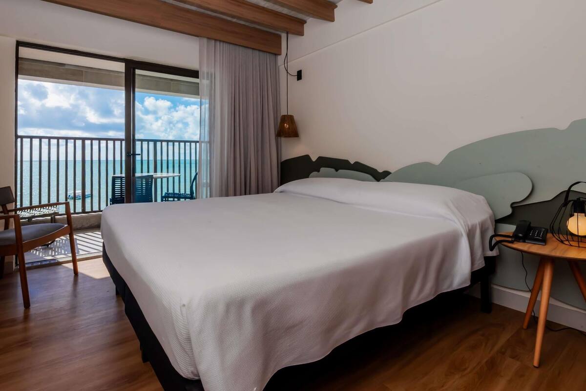 A hotel room with a bed, a chair, a table, a lamp, and a view of the sea.