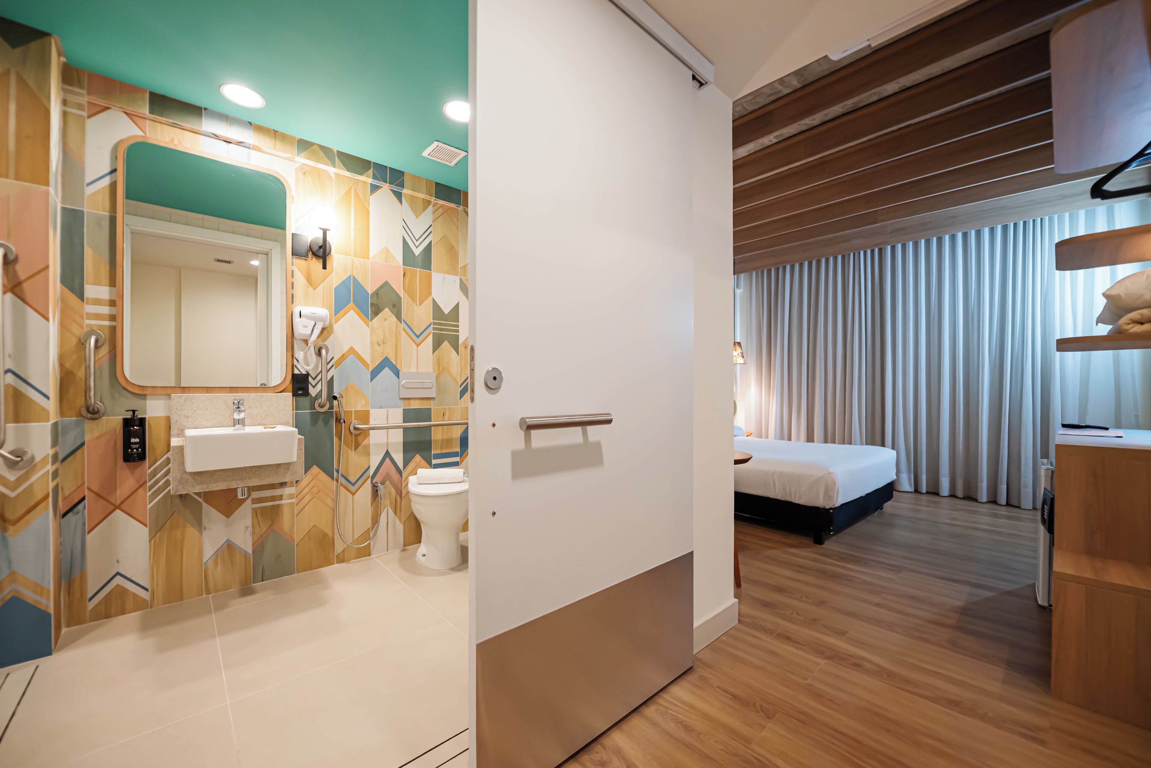 A modern hotel room with a wooden floor, a bed, a bathroom with a mirror, sink, and toilet.