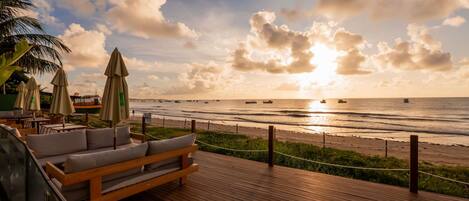 A wooden deck with a sofa and umbrellas overlooking a beach with boats and a sunset.