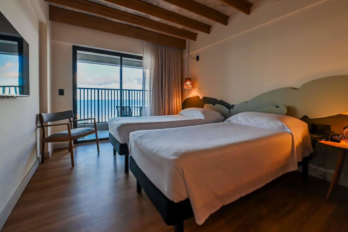 A hotel room with two beds, a chair, a nightstand, and a balcony with a view.