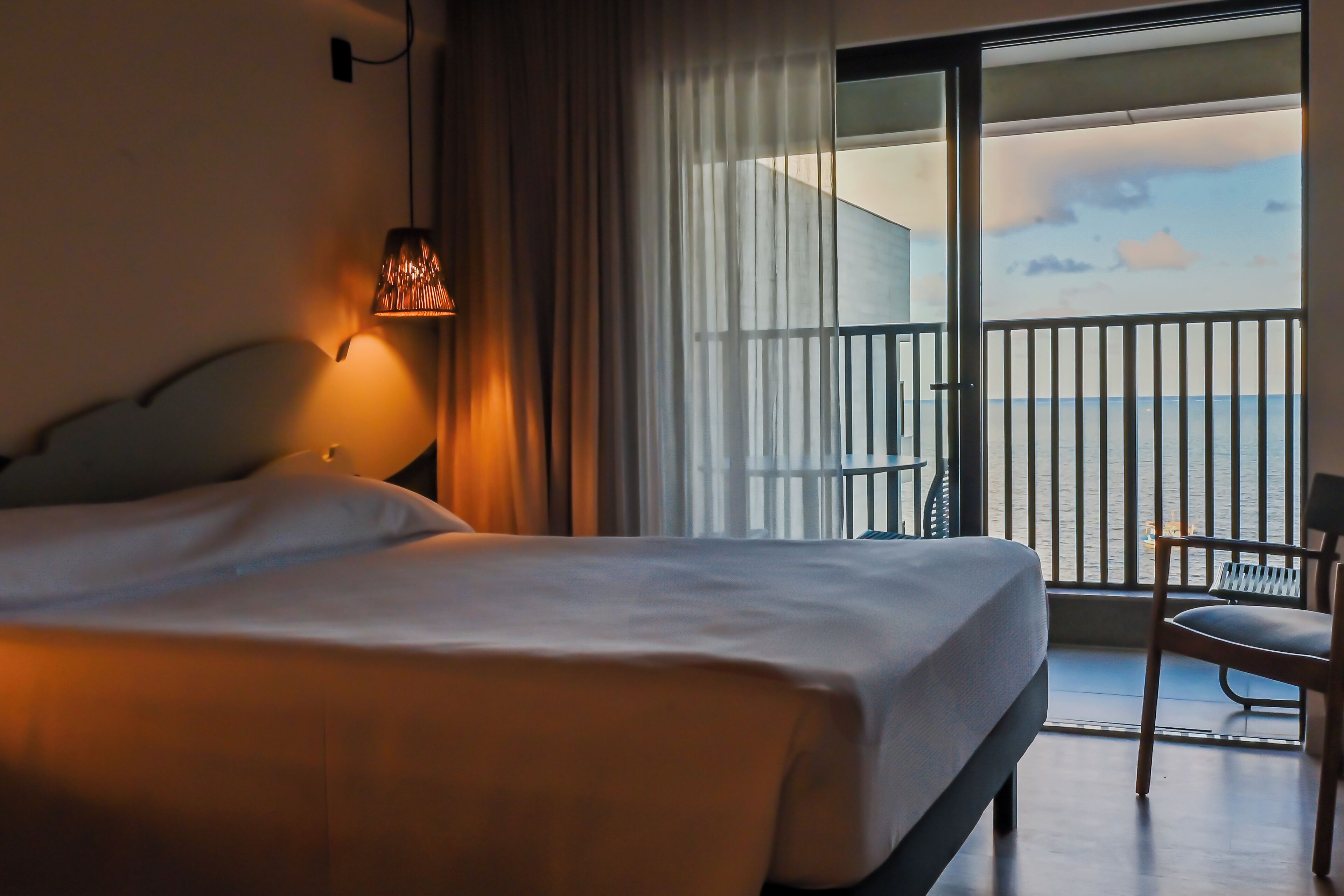 A hotel room with a bed, a chair, a lamp, a balcony with a railing, and a view of the sky.