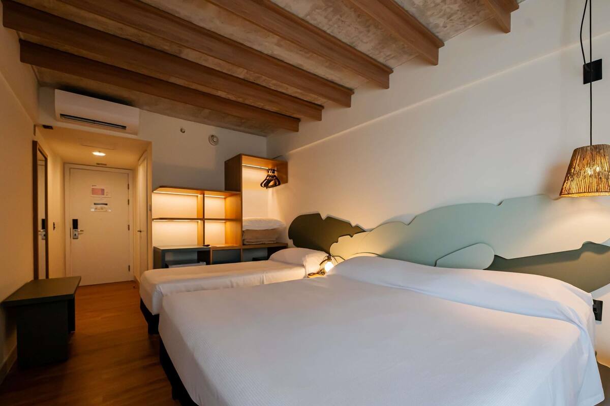A hotel room with a wooden ceiling, a bed with white bedding, a bedside table, a lamp, and a wall-mounted shelf.