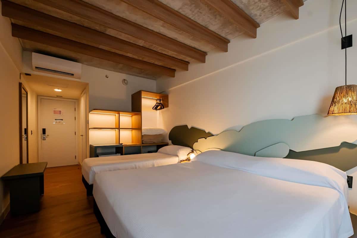 A hotel room with a wooden ceiling, a bed with white bedding, a bedside table, a lamp, and a wall-mounted shelf.