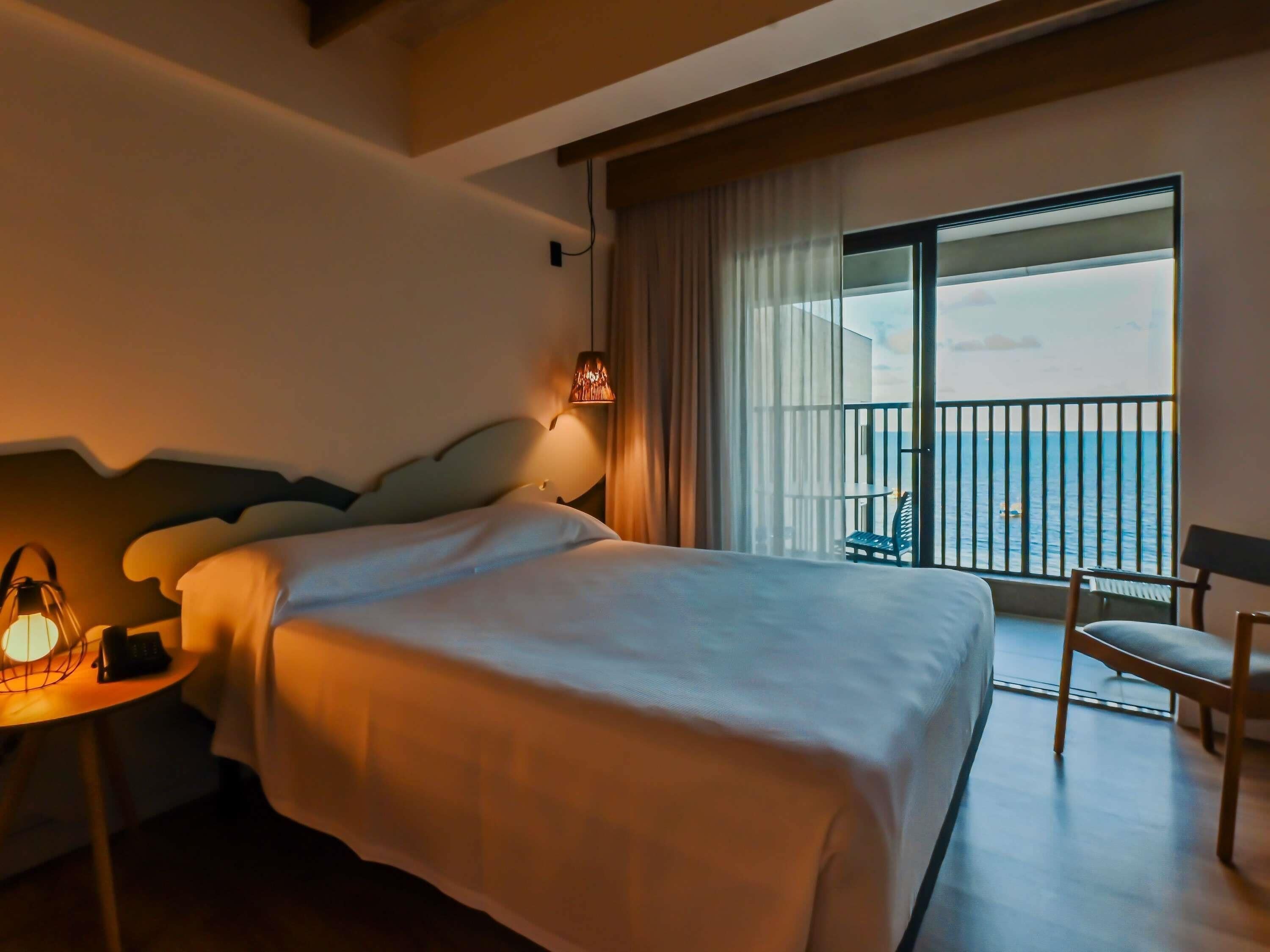 Habitación de hotel con una cama grande, una mesita de noche con lámpara, una silla y un balcón con vistas al mar.