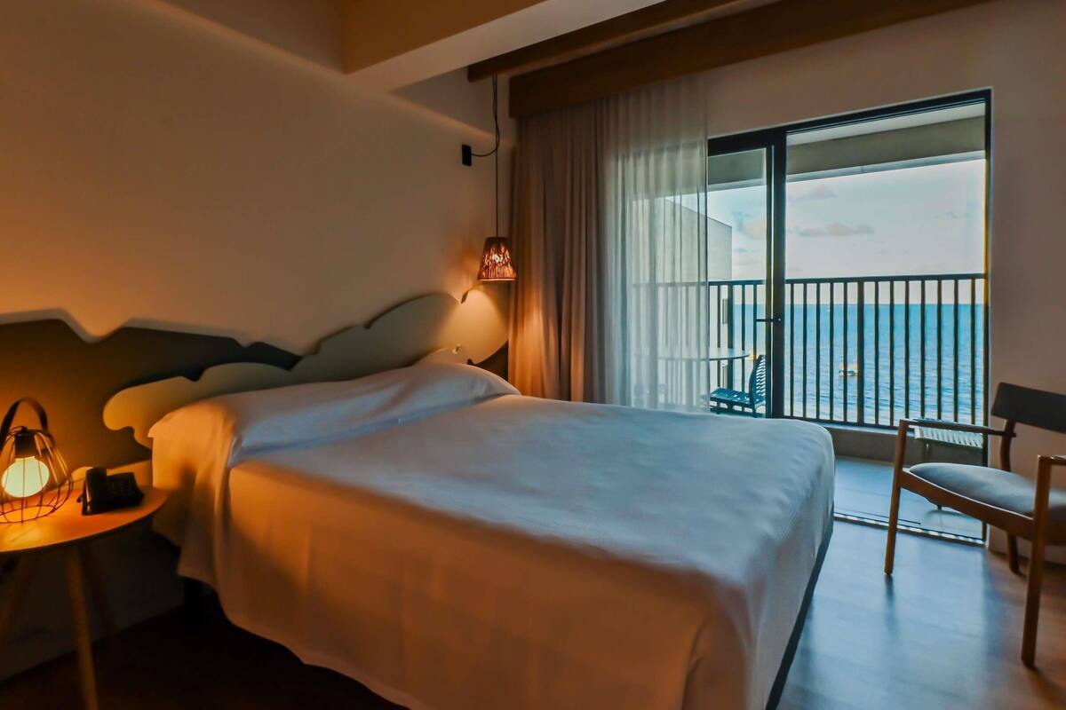 A hotel room with a large bed, a bedside table with a lamp, a chair, and a balcony with a view of the sea.