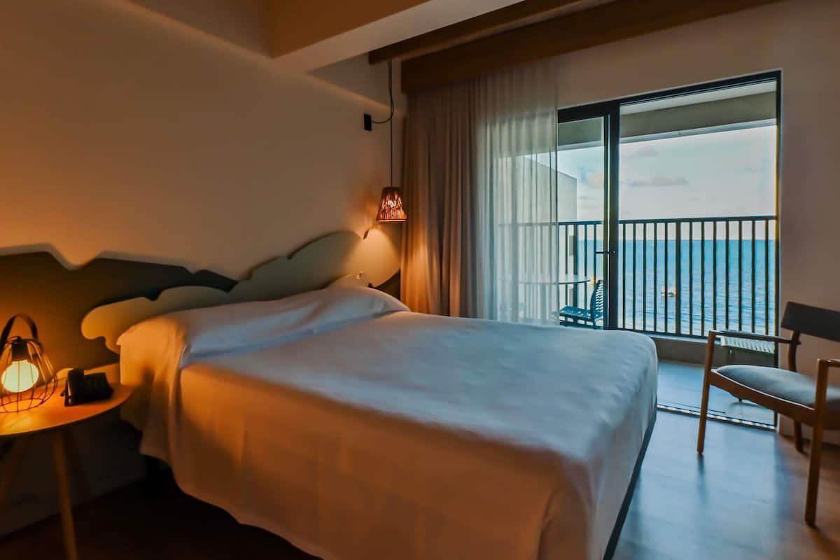 Habitación de hotel con una cama grande, una mesita de noche con lámpara, una silla y un balcón con vistas al mar.