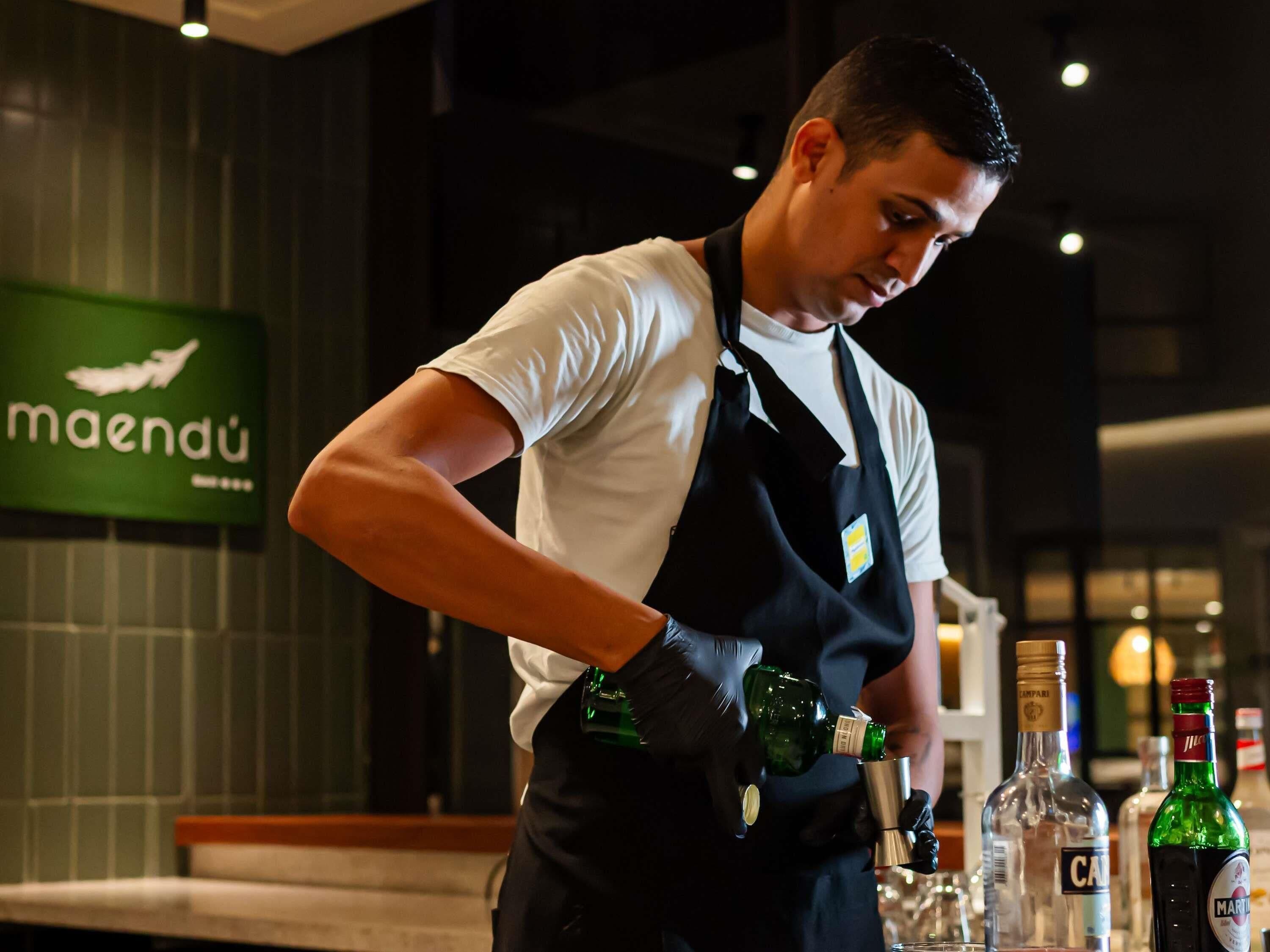 Un barman preparando una bebida en un bar, con un letrero verde al fondo.