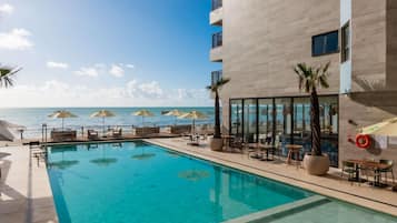 Uma área de piscina com espreguiçadeiras e guarda-sóis, vista desobstruída do oceano e um edifício com amplas janelas.