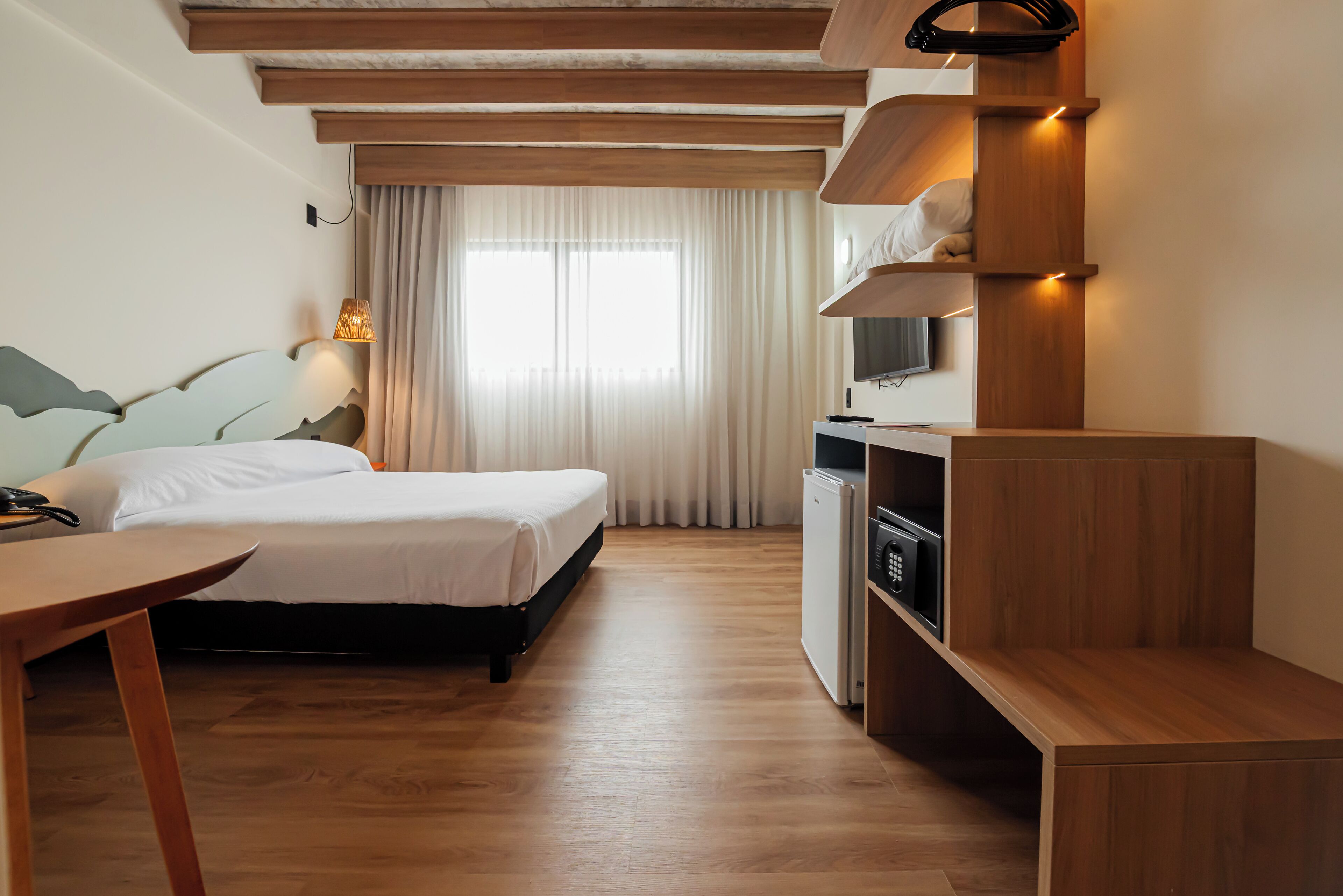 A hotel room with a wooden floor, a bed with white bedding, a desk with a chair, a TV mounted on the wall, and a small kitchenette area.