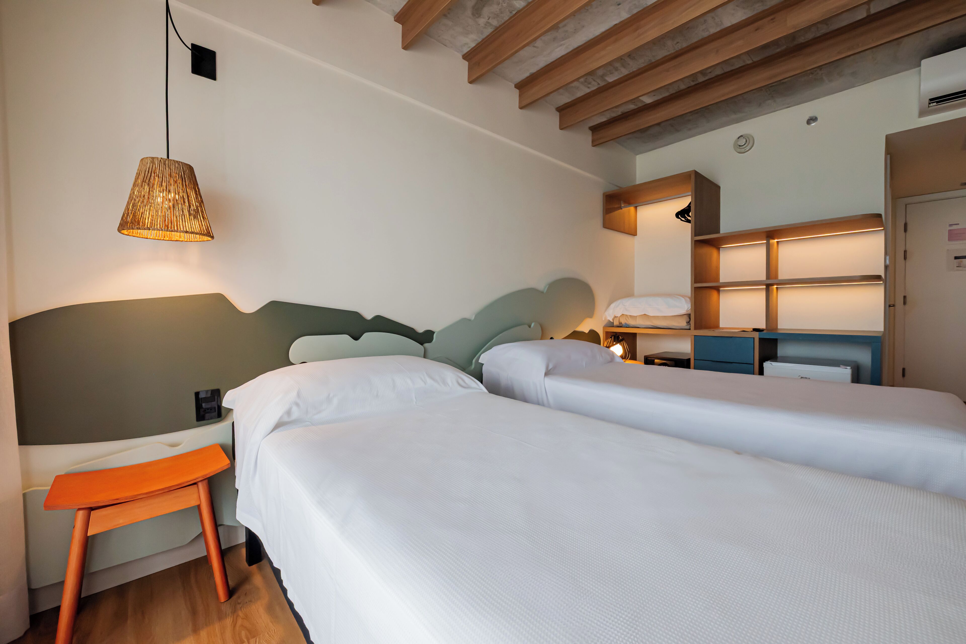A hotel room with two beds, a wooden bench, a hanging lamp, and built-in shelving.