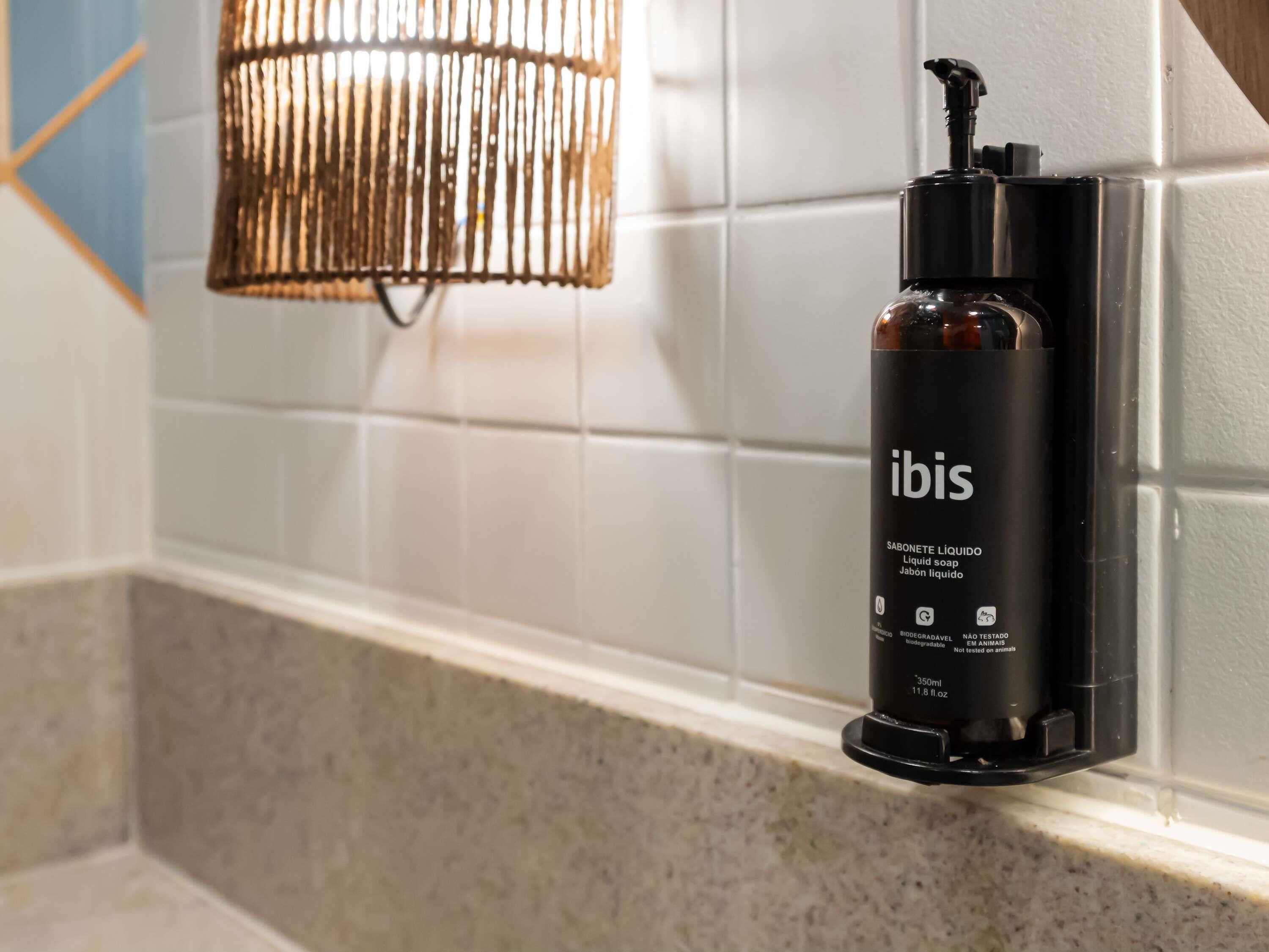Une salle de bain avec un flacon noir contenant du savon liquide, portant l’étiquette « ibis », sur un mur recouvert de carreaux blancs.