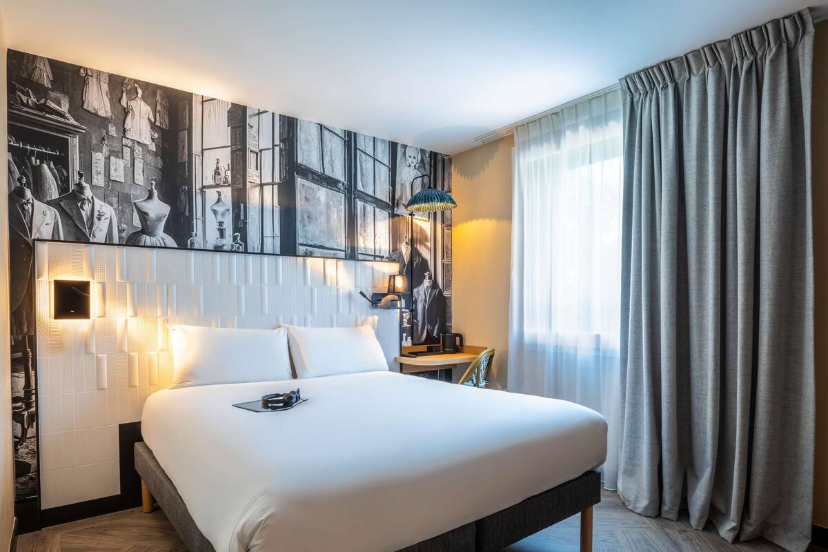 A modern hotel room with a large bed, a desk, and a wall with a cityscape mural.