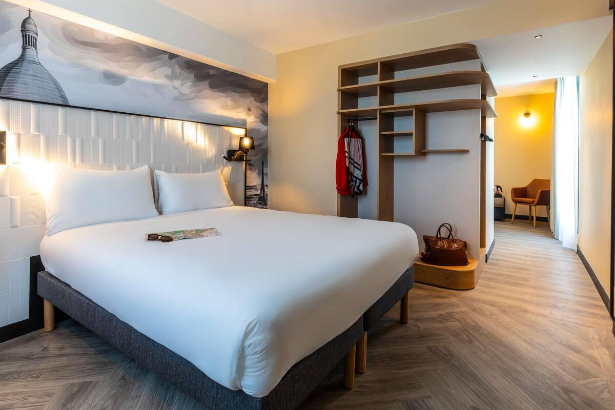 A modern hotel room with a large bed, a headboard with a cityscape mural, a wooden sideboard, and a view of the city through the window.