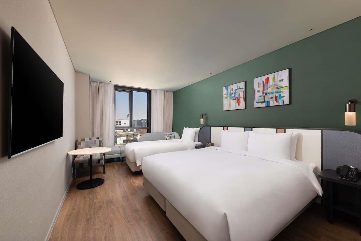 A hotel room with two beds, a TV, a desk, and a window with a city view.