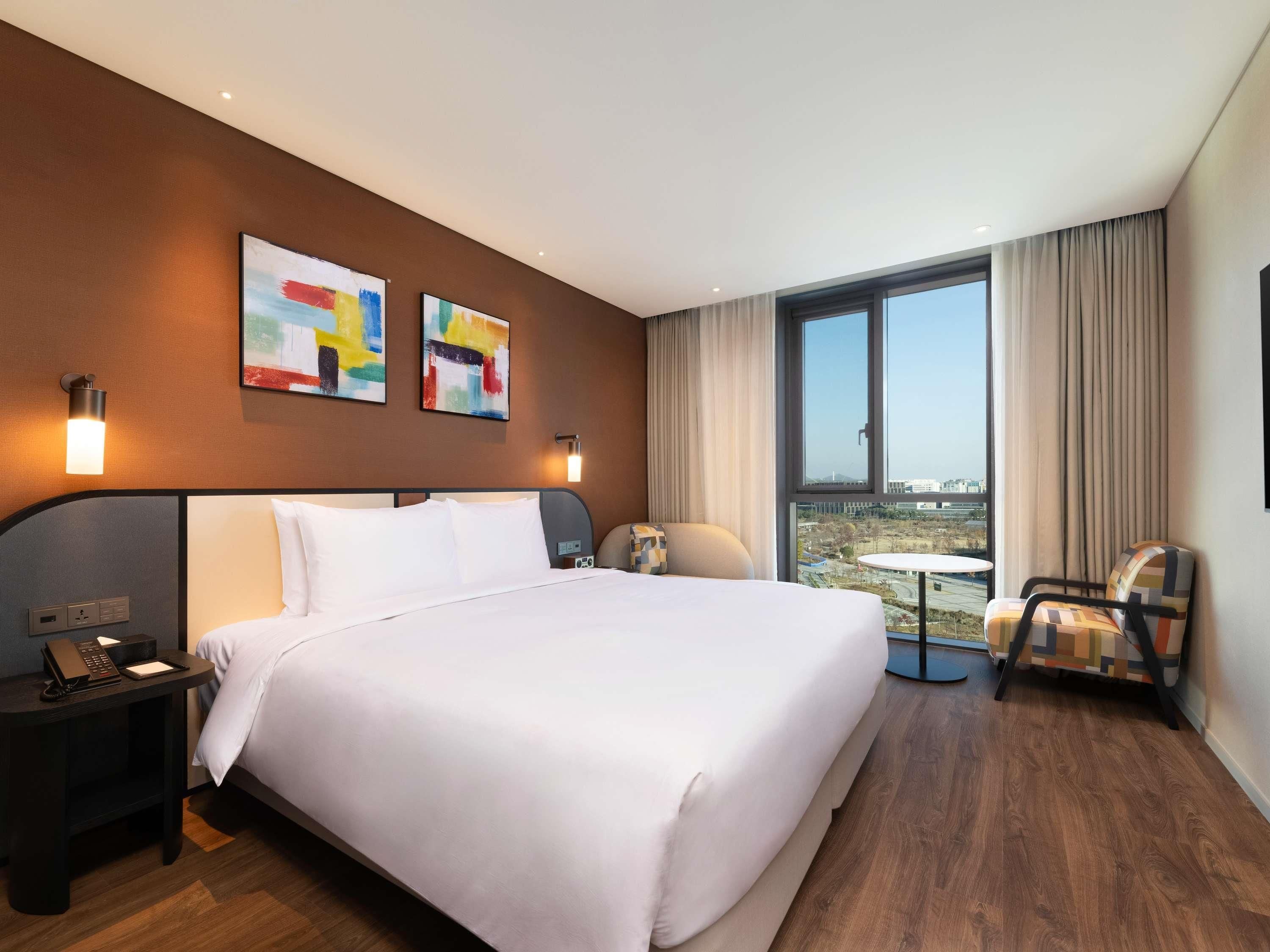 A hotel room with a large bed, a desk with a phone, two wall-mounted lamps, a chair, and a view of the city through a large window.