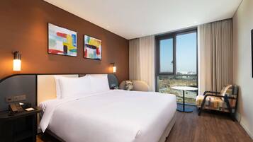 A hotel room with a large bed, a desk with a phone, two wall-mounted lamps, a chair, and a view of the city through a large window.